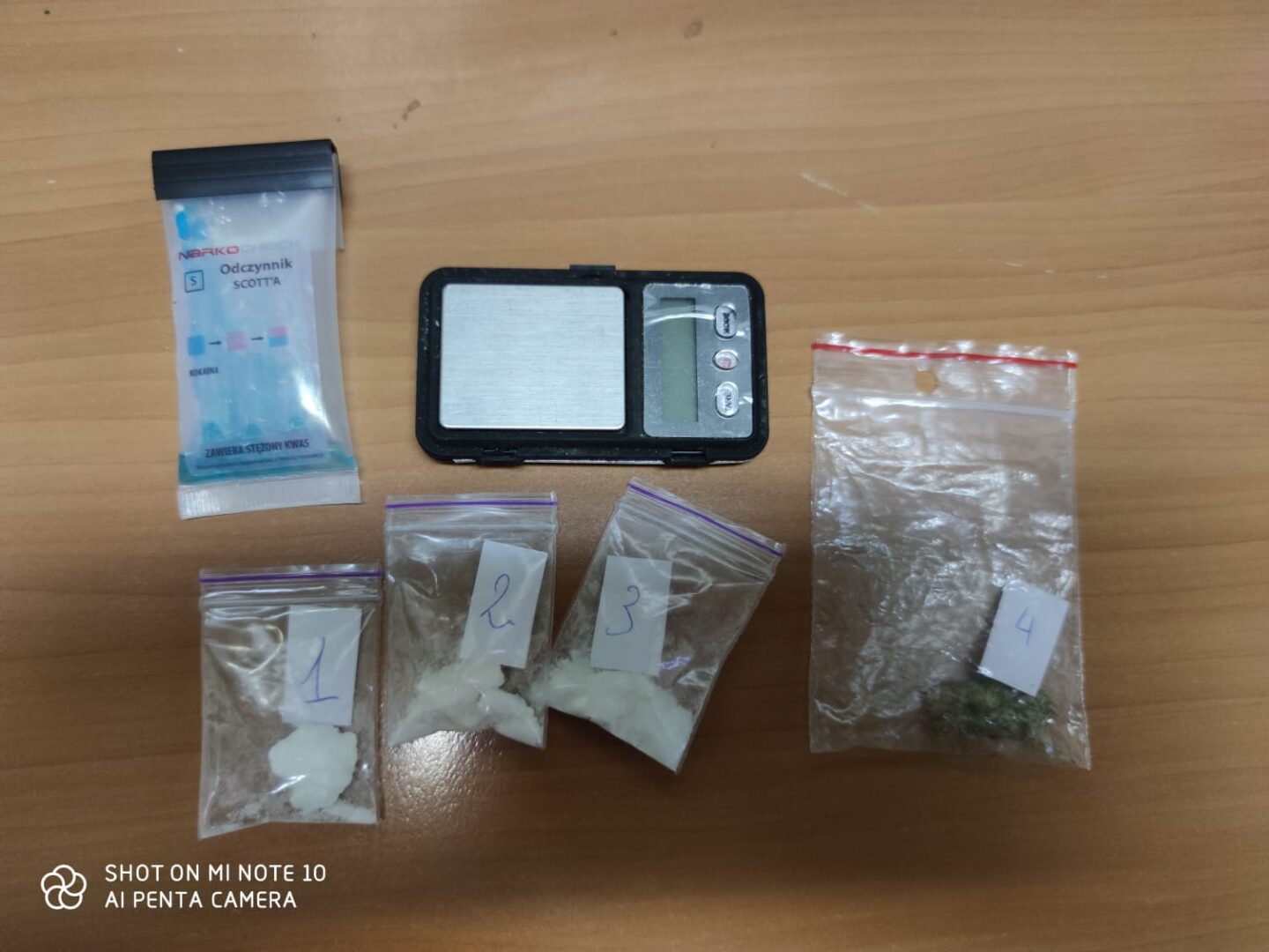 Cztery torebki z białym proszkiem i jedna torebka z zielonym materiałem roślinnym, obok wagi cyfrowej i zestawu do testów narkotykowych.