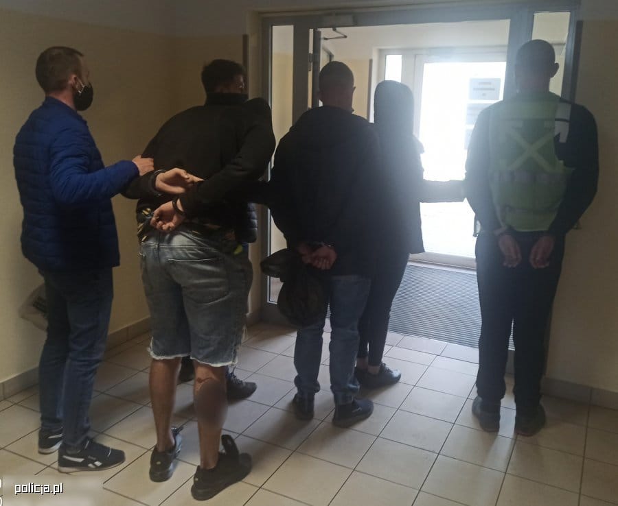 Czterech podejrzanych w kajdankach, eskortowanych przez funkcjonariuszy policji.