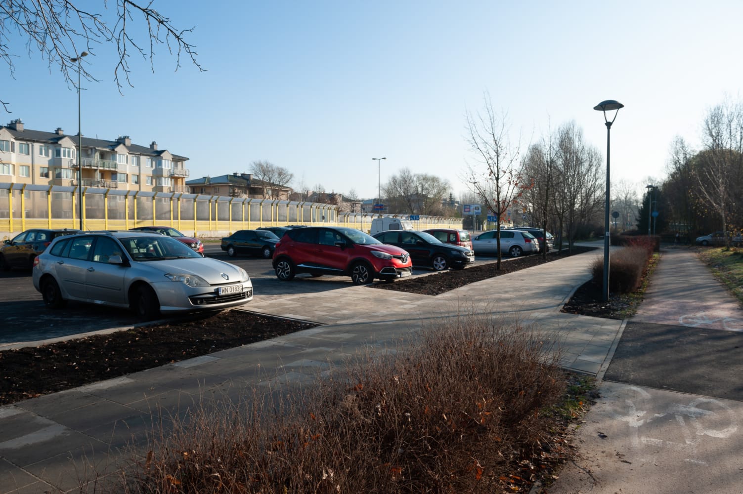 Samochody zaparkowane na parkingu w pobliżu budynku mieszkalnego i ścieżki rowerowej.