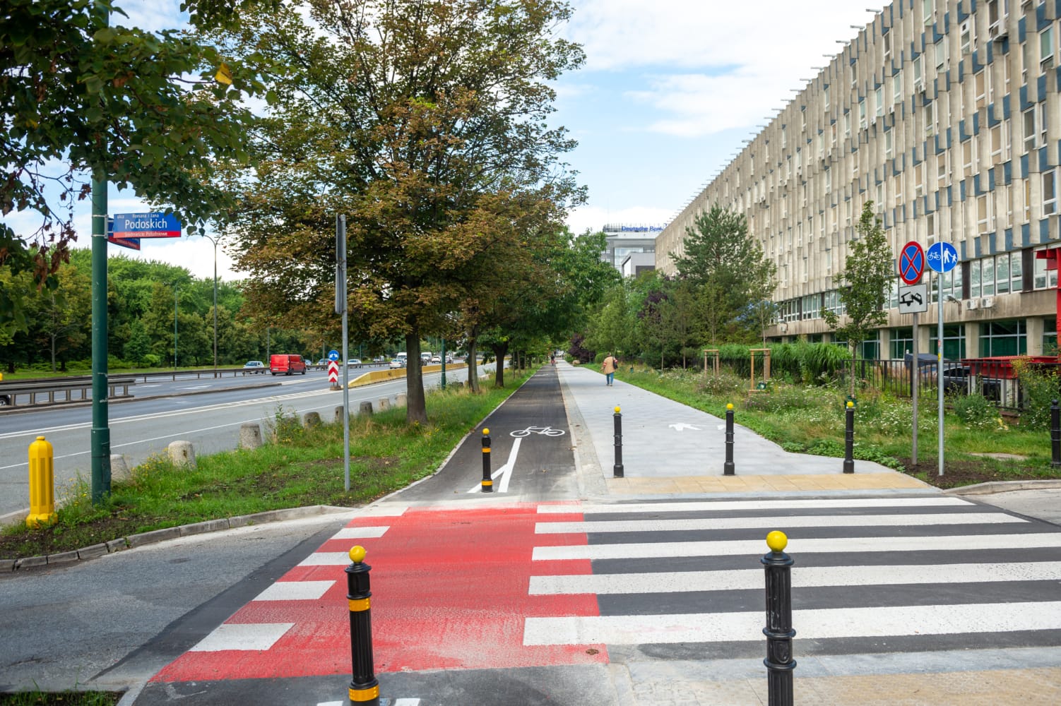 Przejście dla pieszych z pasem rowerowym i nowoczesnym budynkiem w tle. Widoczny znak uliczny "Podoskich".