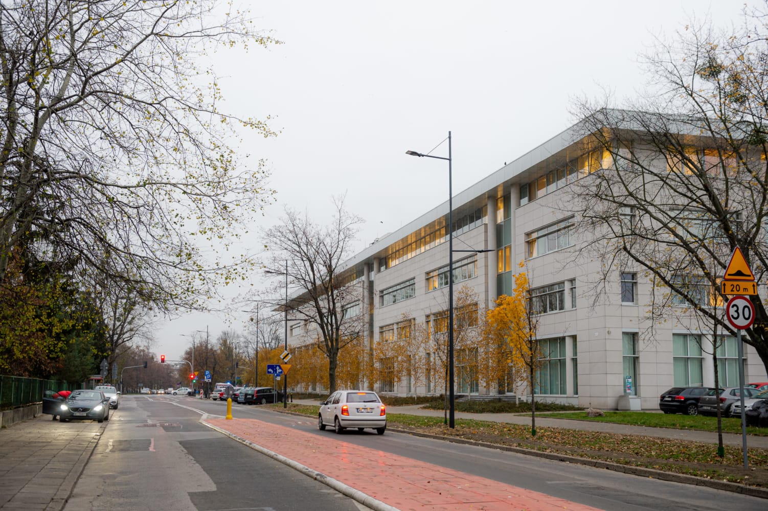 Nowoczesny budynek biurowy na miejskiej ulicy, wśród jesiennych drzew i samochodów.