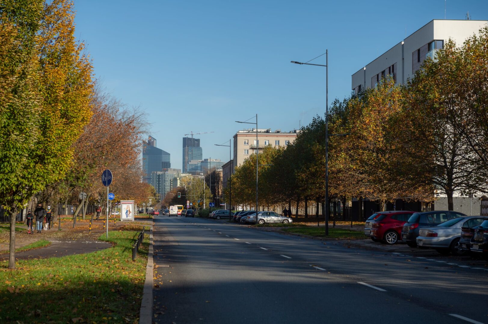 Jesienny widok na ulicę miasta, zaparkowane samochody i nowoczesne wieżowce w oddali.