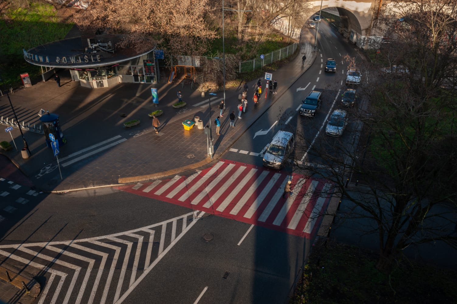 Widok z lotu ptaka na skrzyżowanie w mieście z pieszymi i samochodami, widocznym przejściem dla pieszych i znakiem „Stacja Powiśle”.