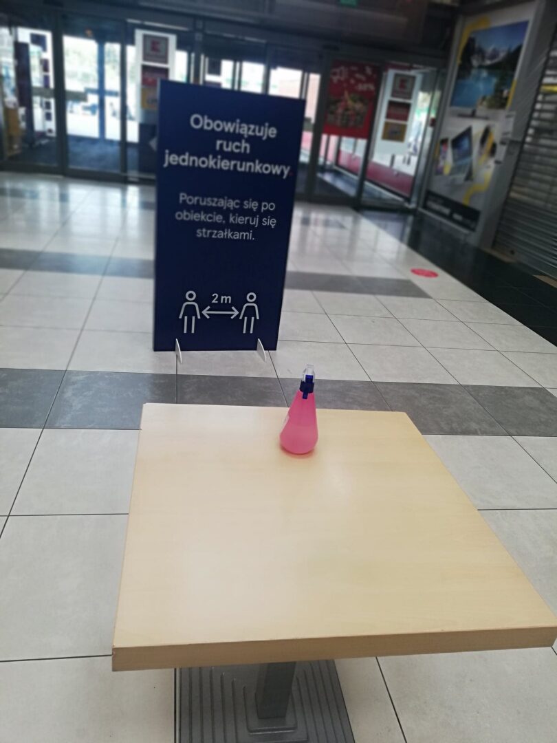 Znak na podłodze wskazujący ruch jednokierunkowy z zachowaniem 2-metrowego dystansu, środek do dezynfekcji rąk na stole