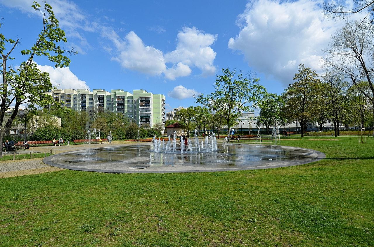 Dzieci bawią się w okrągłej fontannie w parku niedaleko budynków mieszkalnych w słoneczny dzień.