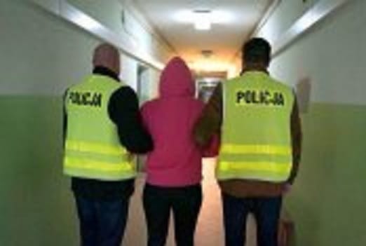 Dwóch policjantów eskortuje kobietę korytarzem. Na ich kamizelkach widać napis POLICJA.