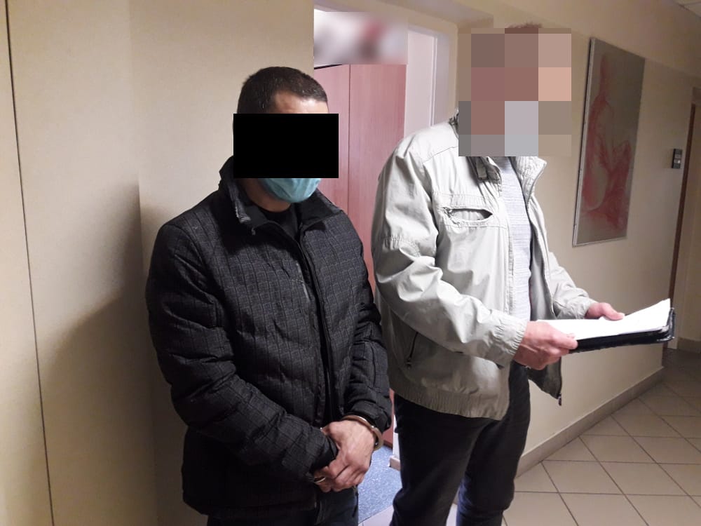 Dwóch mężczyzn w kajdankach, jeden trzymający dokumenty, podczas aresztowania.