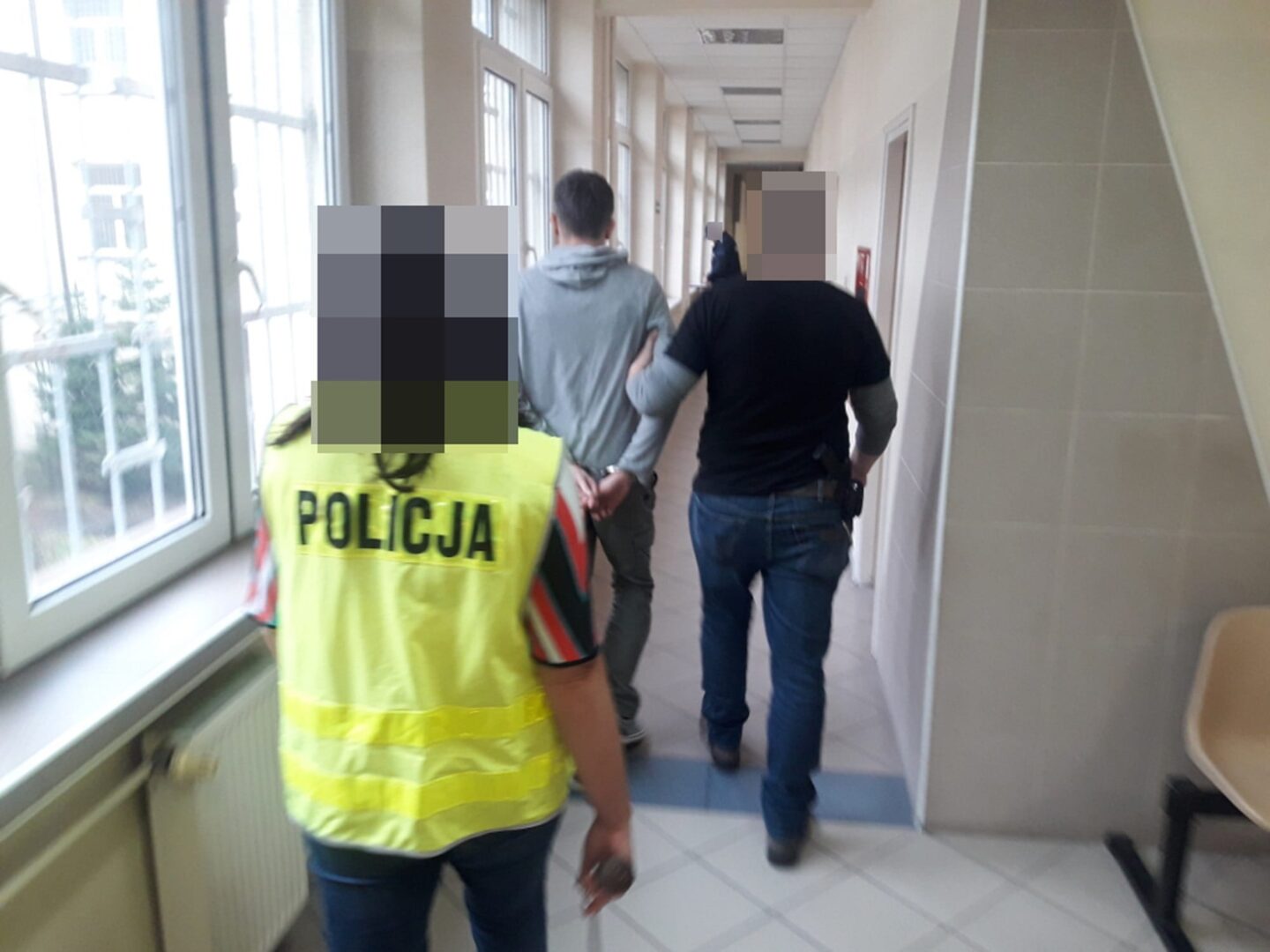 Policja eskortuje podejrzanego korytarzem. Na kamizelce oficera widać napis „Policja”.