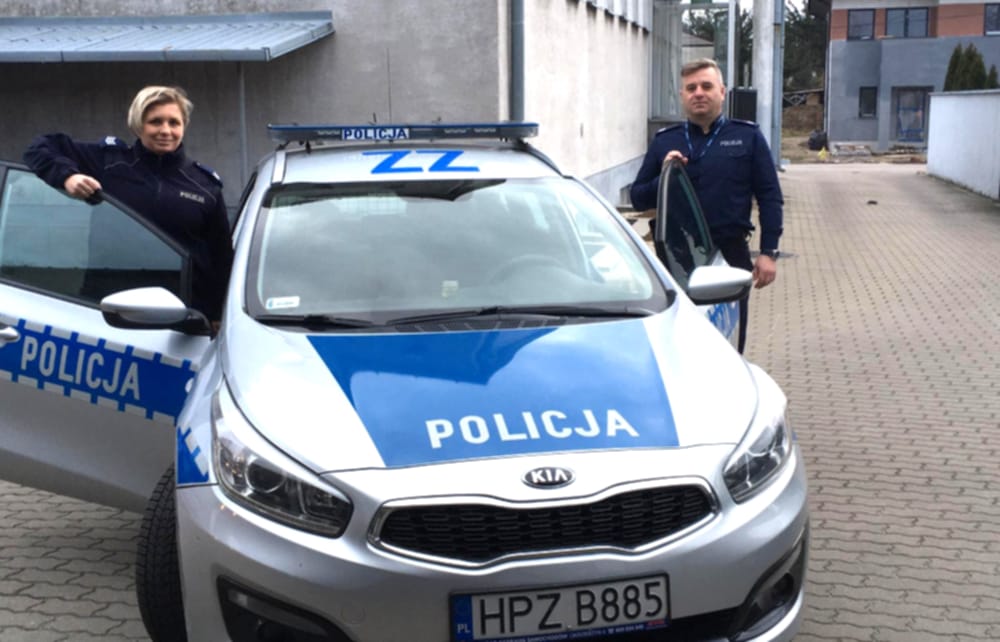 Dwóch polskich policjantów stoi dumnie przy swoim radiowozie z napisem „POLICJA”.