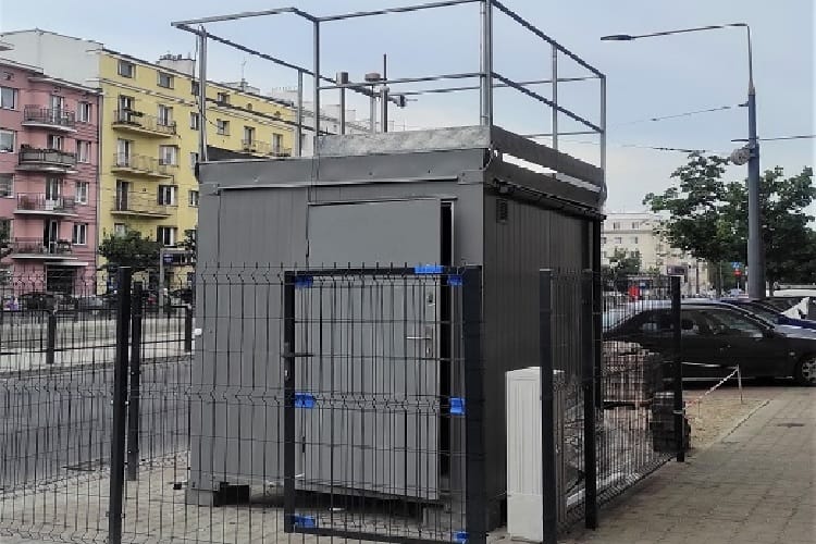 Nowoczesna, szara publiczna toaleta z ogrodzeniem zabezpieczającym i dostępem z dachu.