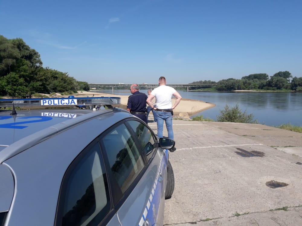 Polscy policjanci prowadzą dochodzenie w pobliżu rzeki; na radiowozie widoczny jest napis „Policja”.