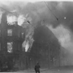 Płonący budynek w czasie wojny, konstrukcja ogarnięta dymem i płomieniami, żołnierz obserwujący scenę.