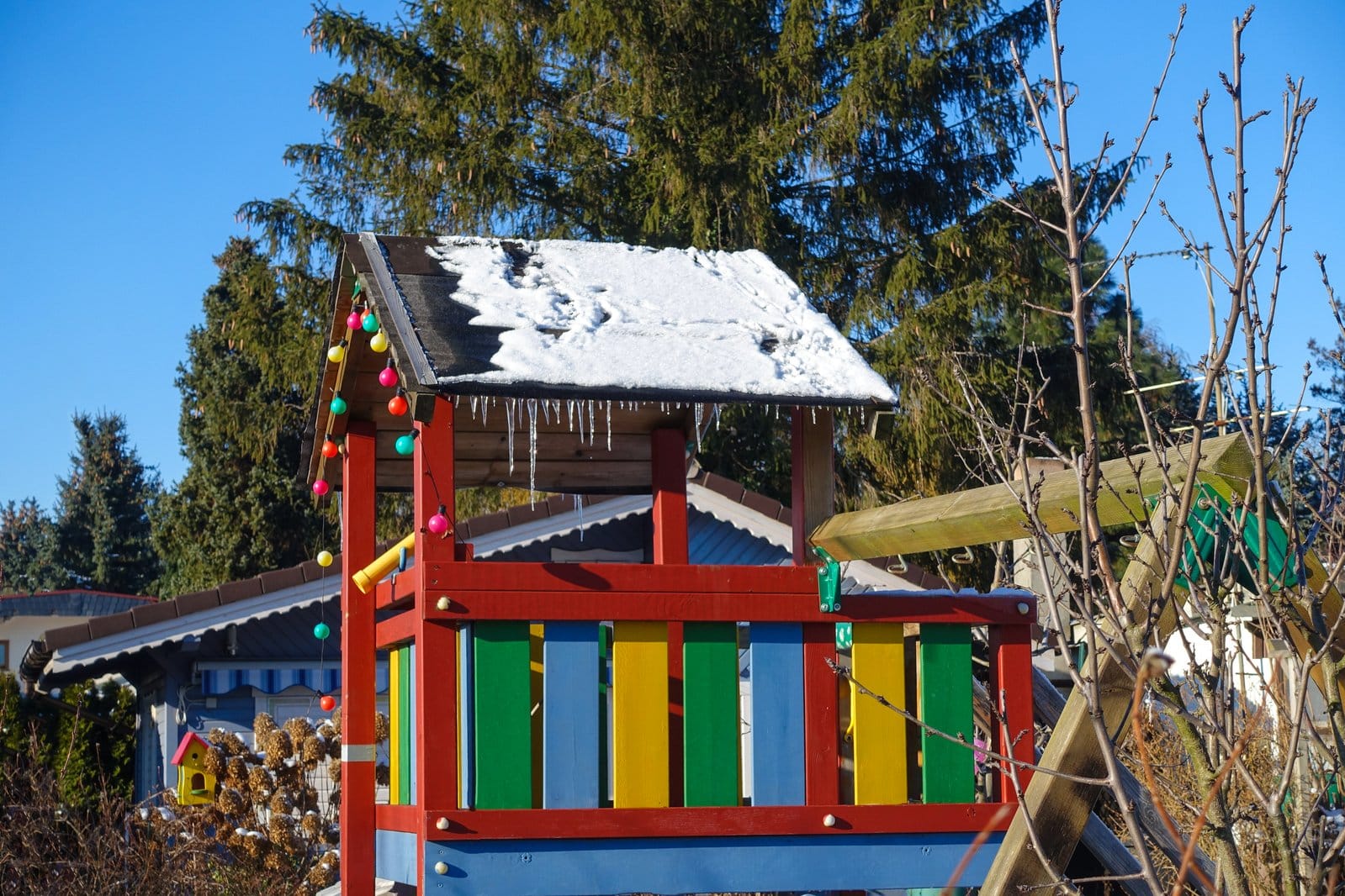 Kolorowy, drewniany plac zabaw pokryty śniegiem, z dachem zwisają sople.