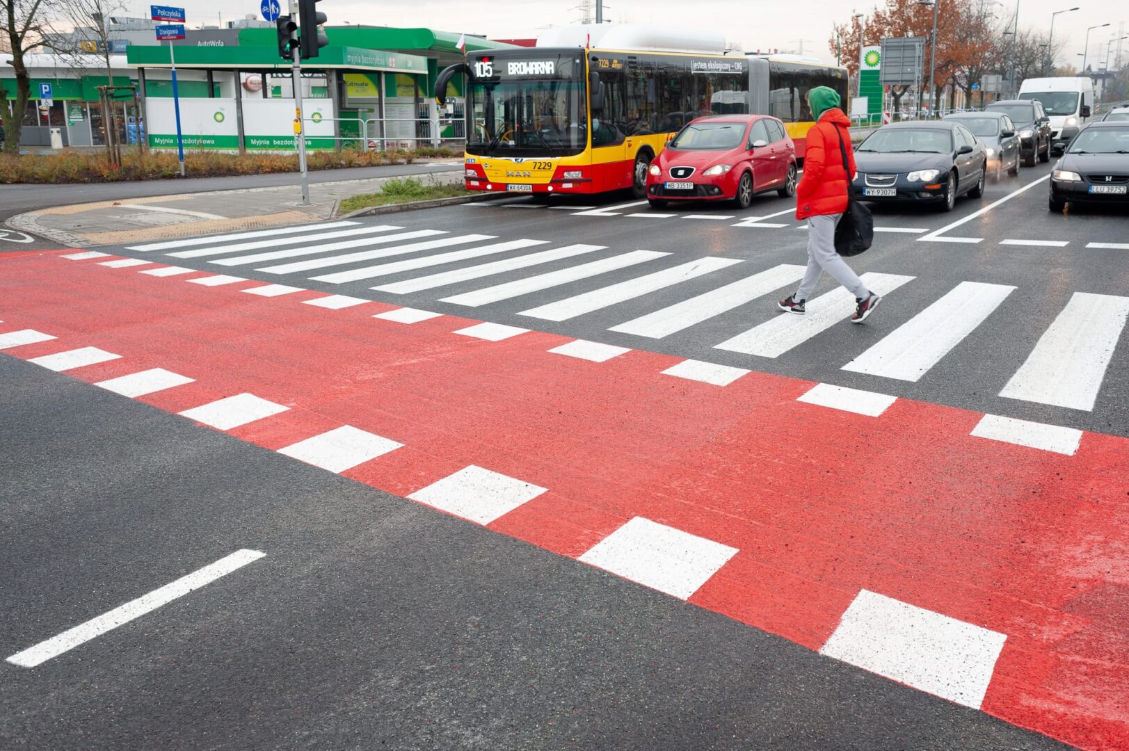 Osoba przechodząca przez przejście dla pieszych oznaczone czerwonymi i białymi znakami, w pobliżu autobusu i samochodów.