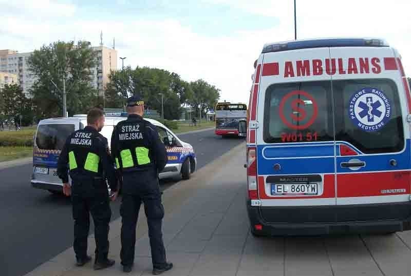 Dwóch polskich strażników miejskich stoi przy karetce i radiowozie. Na ich mundurach widać napis „Straż Miejska”.