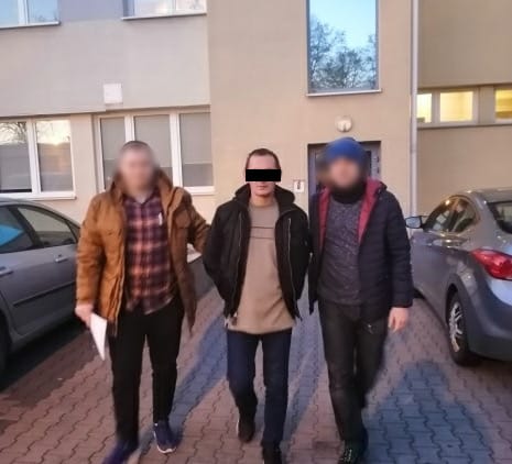 Trzech mężczyzn stoi przed budynkiem, prawdopodobnie po aresztowaniu. Jeden mężczyzna jest w środku, otoczony przez dwóch innych.