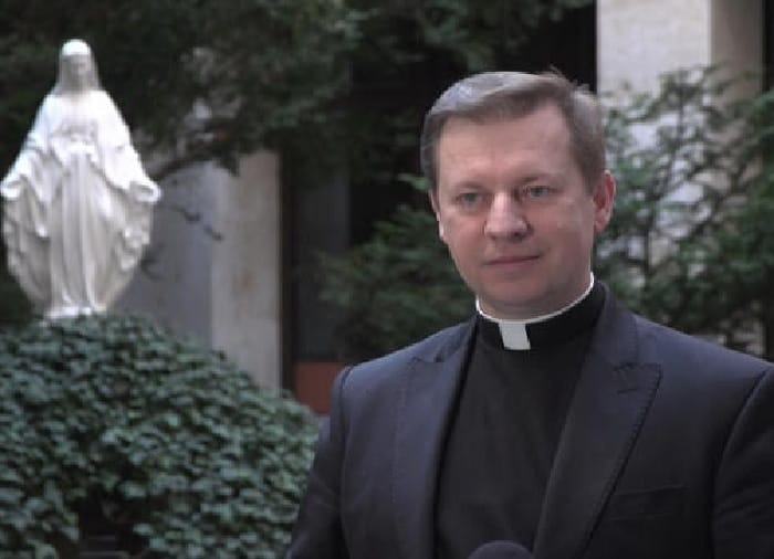 Ksiądz w czarnym koloratce przemawia, w tle widać figurę Maryi