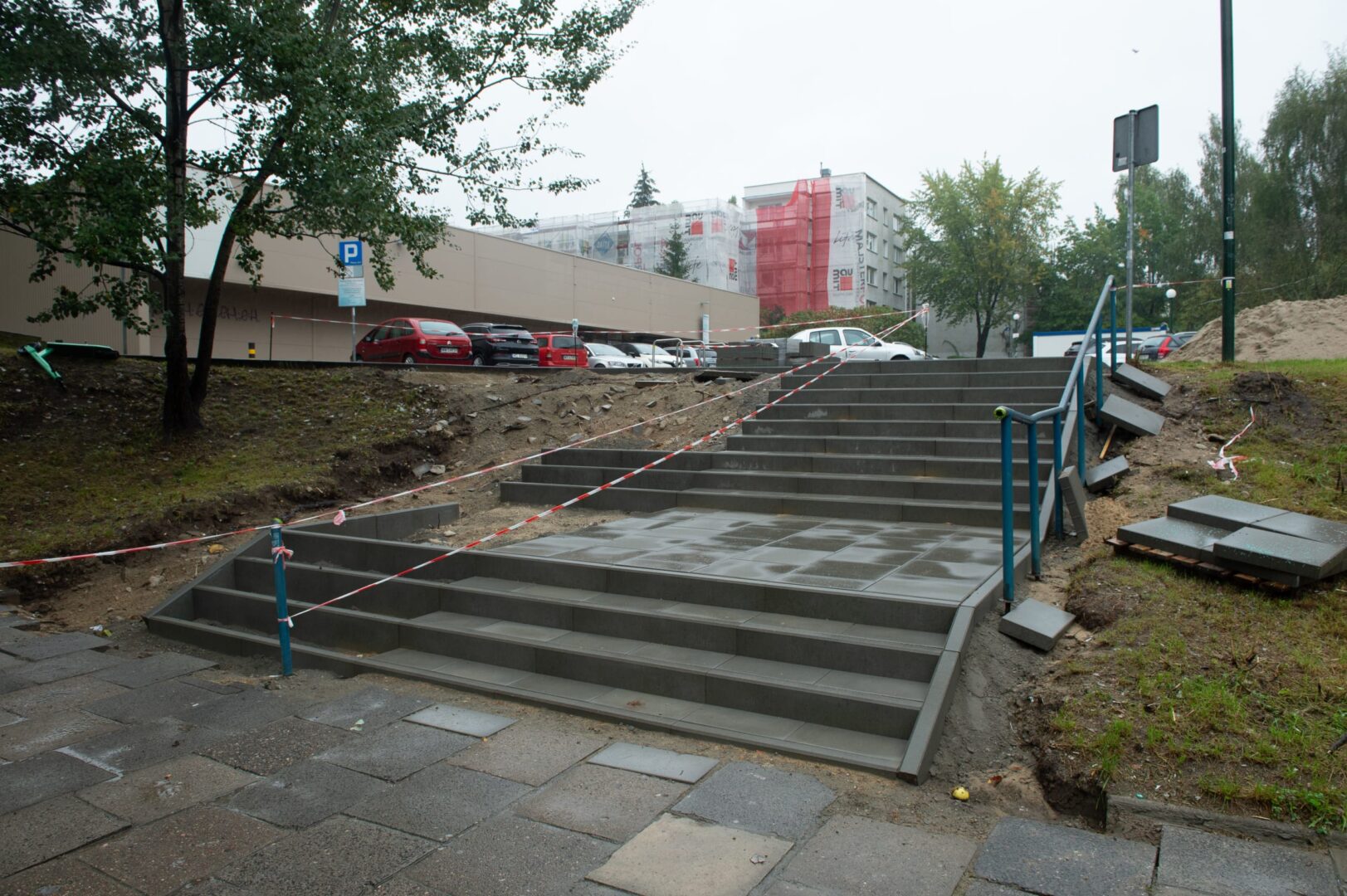 Nowo wybudowane betonowe schody i chodnik w budowie, częściowo ogrodzone czerwoną i białą taśmą ostrzegawczą.