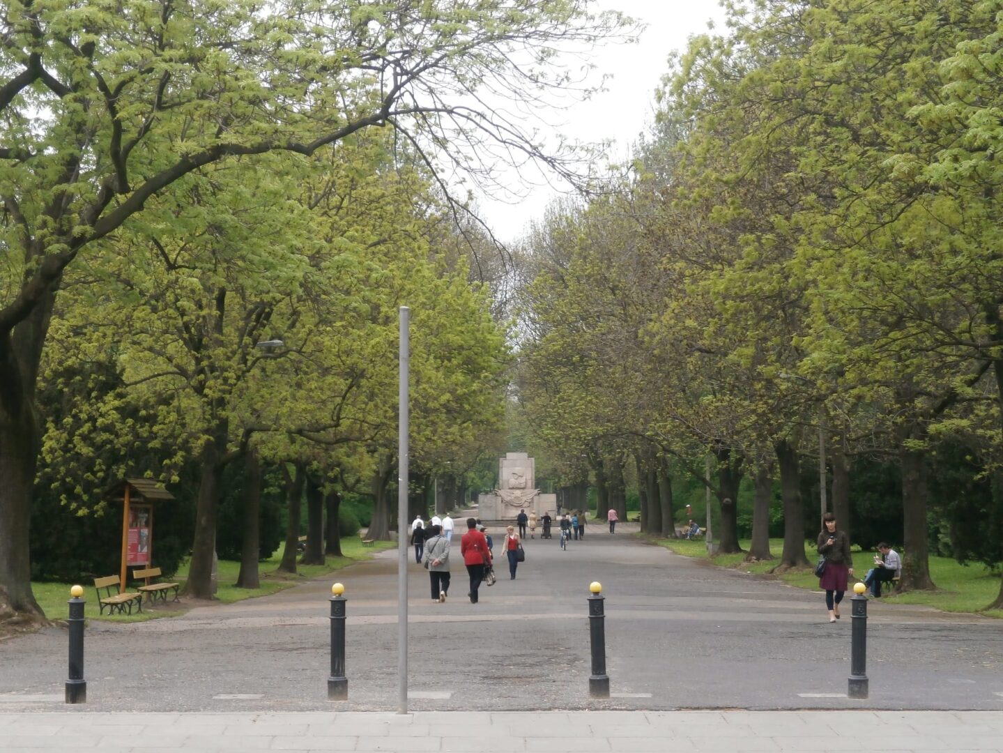Ludzie spacerują aleją wysadzaną drzewami w Parku Skaryszewskim w kierunku pomnika.