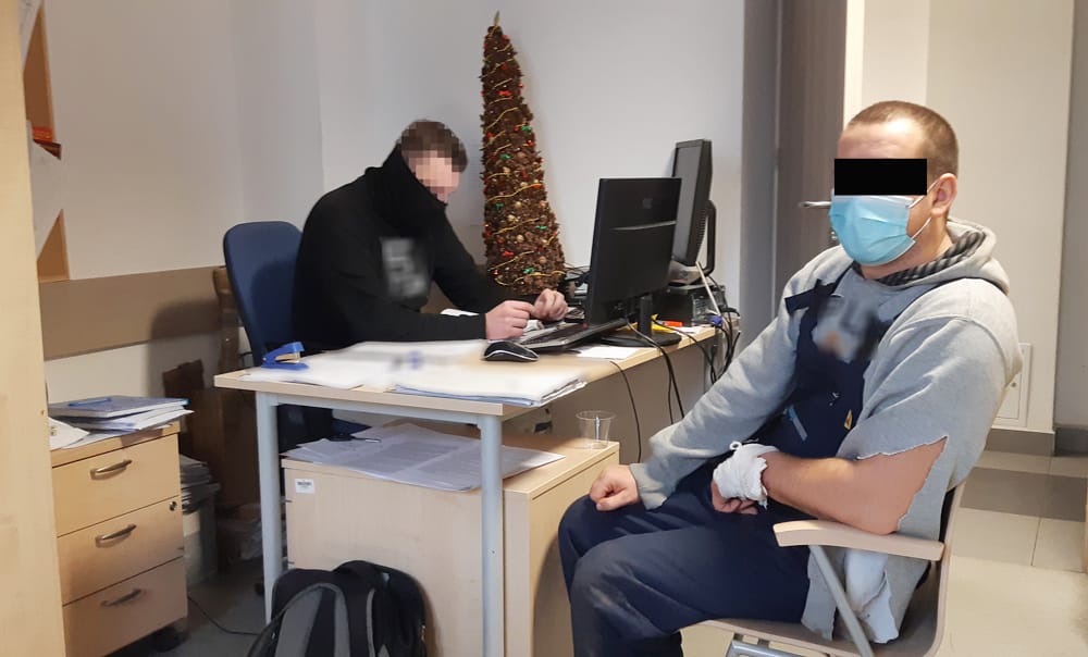 Dwóch mężczyzn w biurze, jeden pracuje przy komputerze, drugi ma bandaż na ramieniu i maskę.