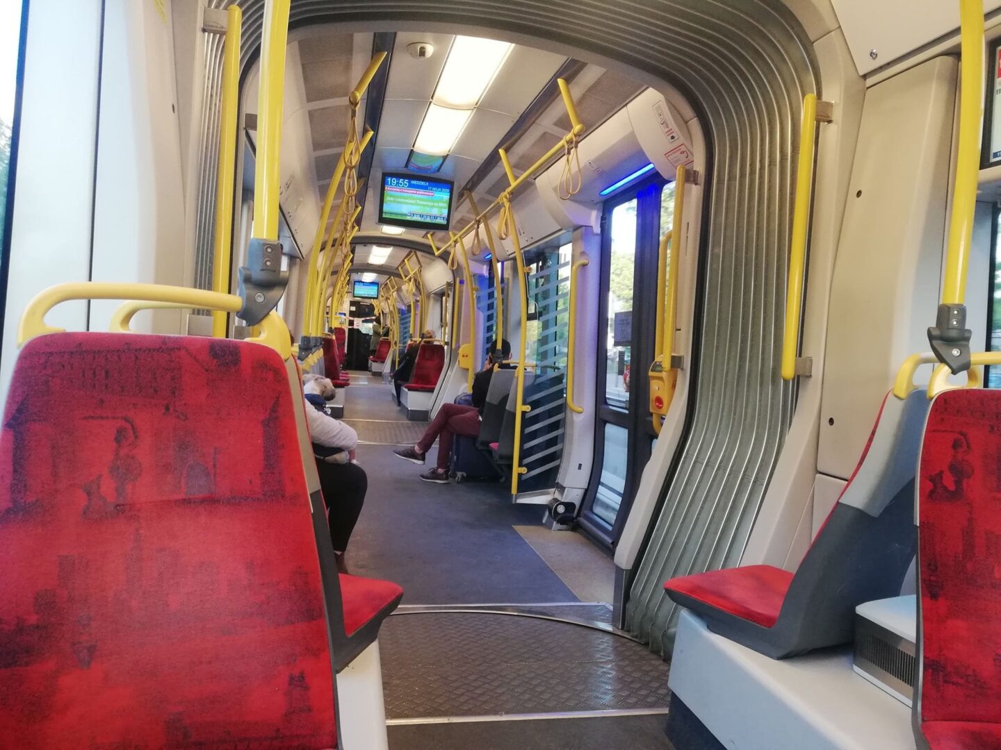 Puste siedzenia w nowoczesnym tramwaju, z czerwoną tapicerką i żółtymi poręczami. Cyfrowy wyświetlacz pokazuje godzinę.