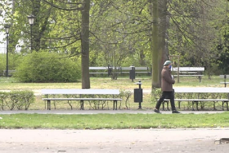 Osoba w masce idzie przez park z ławkami i drzewami.