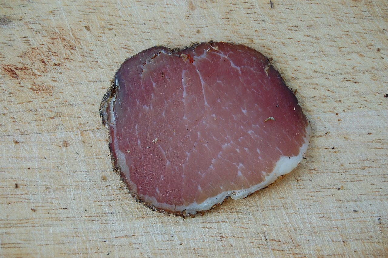 Zbliżenie pojedynczego plasterka wędzonej polędwicy wieprzowej położonego na drewnianej desce do krojenia.