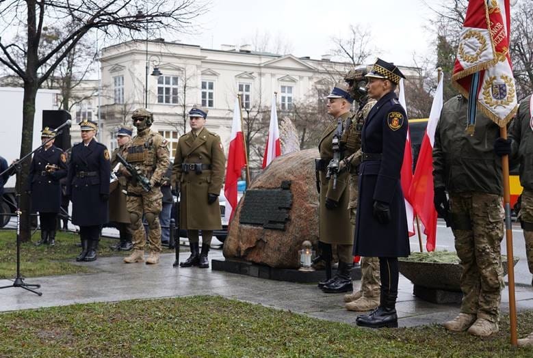 Polscy oficerowie i policjanci stoją w szyku przy pomniku, upamiętniającym rocznicę zamachu.