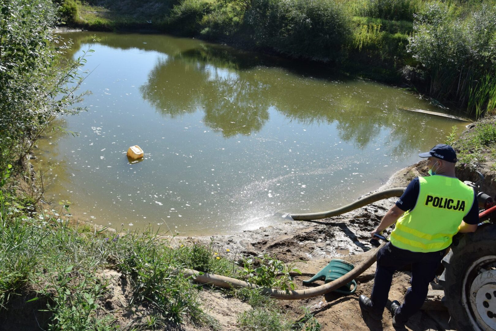 Policjant pompuje skażoną wodę ze stawu. Na kamizelce policjanta widoczny jest napis „POLICJA”.