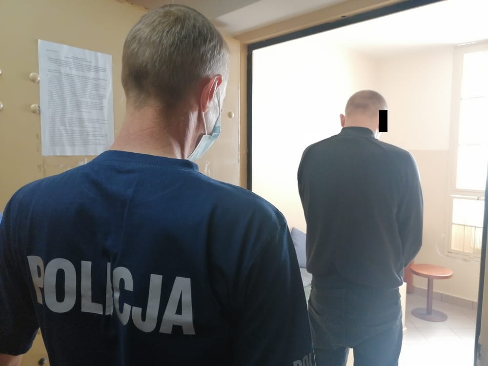 Policjant z napisem „POLICJA” na plecach, stojący za podejrzanym w pokoju.