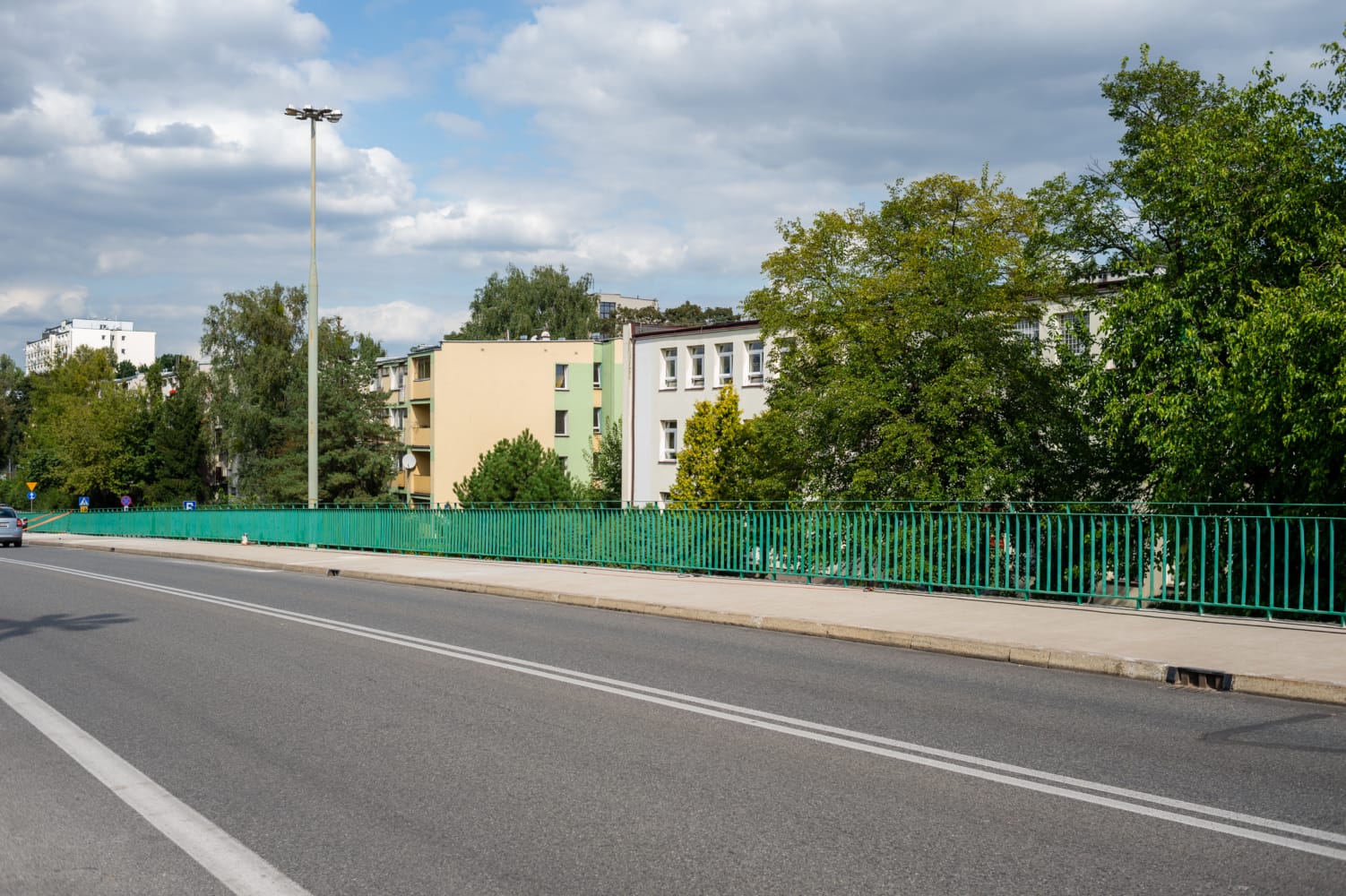 Nowoczesne budynki mieszkalne wzdłuż drogi miejskiej z zielonym płotem.