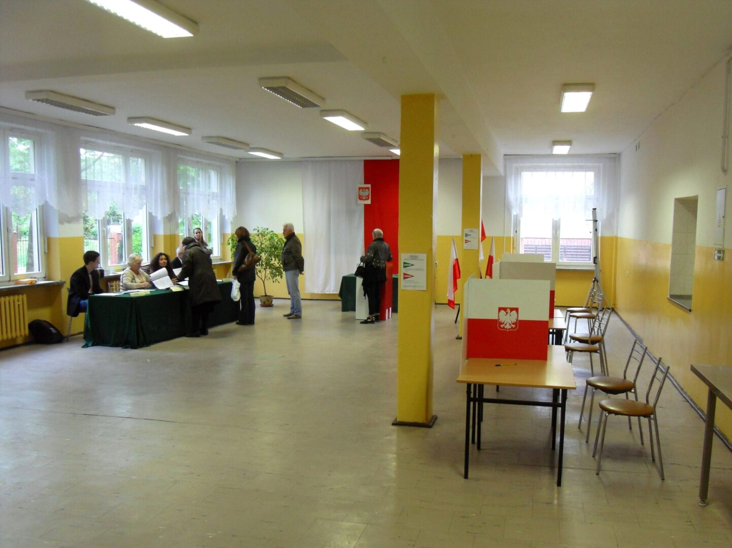 Wnętrze lokalu wyborczego z wyborcami i urzędnikami wyborczymi. Widoczne są polskie flagi i kabiny do głosowania.