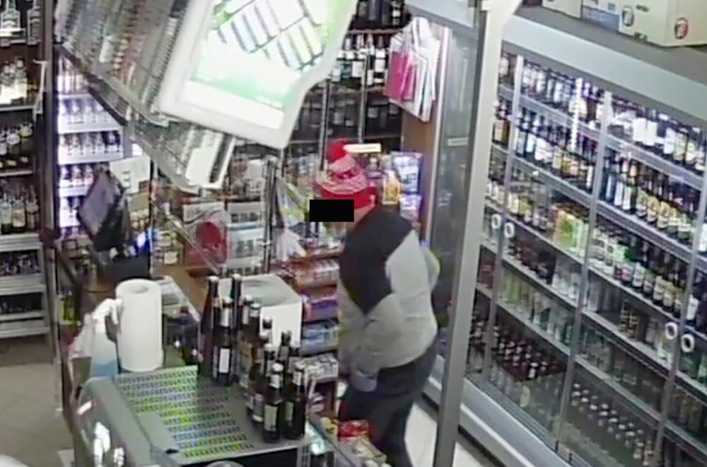 Obraz z monitoringu przedstawiający złodzieja sklepowego w sklepie spożywczym, ubranego w szary sweter i świąteczną czapkę. Złodziej znajduje się przy przewróconym monitorze komputera.