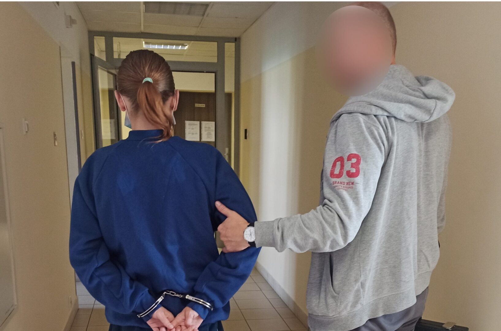 Policjant eskortujący skutą kajdankami kobietę korytarzem.