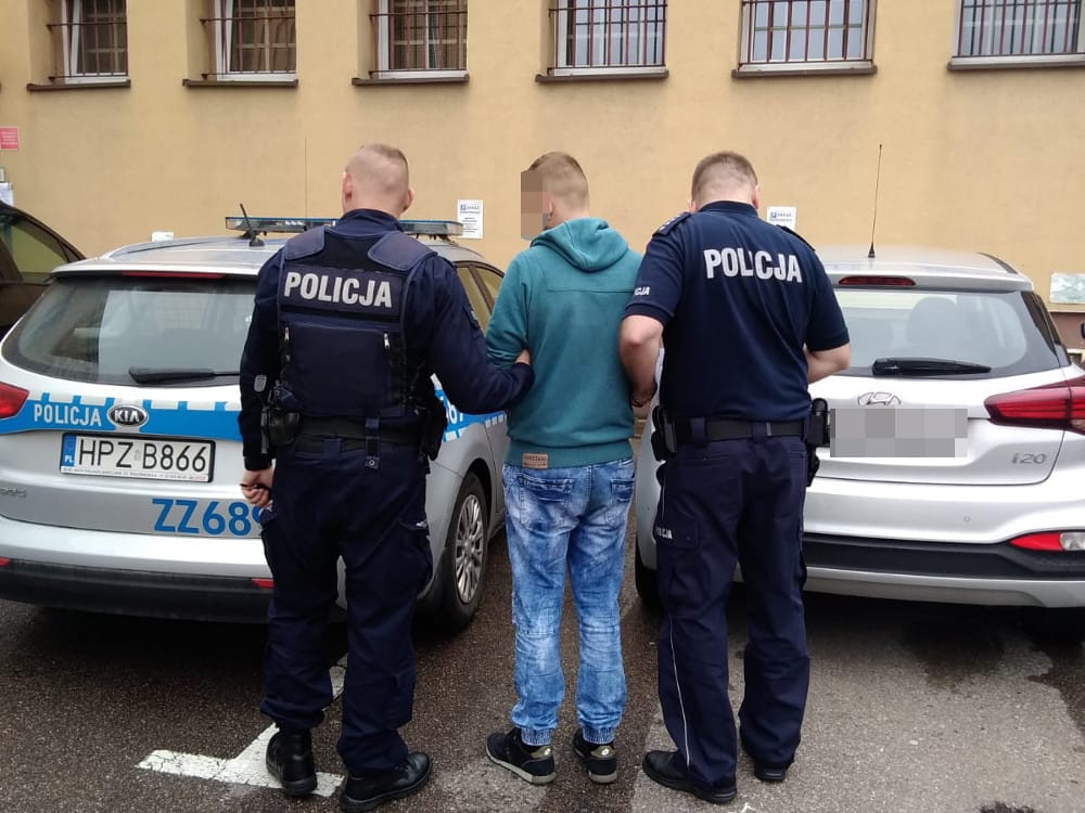 Dwóch polskich policjantów aresztuje podejrzanego pomiędzy dwoma radiowozami. Na mundurach policjantów widać napis „Policja”.