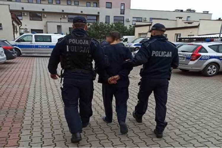 Dwóch polskich policjantów eskortuje podejrzanego w kajdankach. Na ich mundurach widać napis POLICJA.