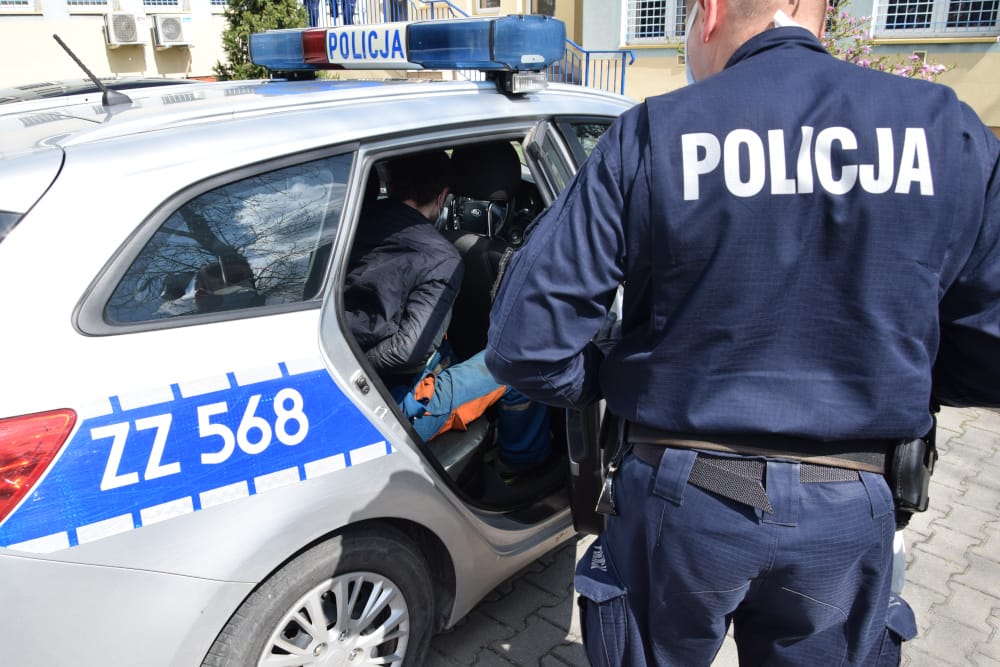 Polski policjant aresztujący podejrzanego w radiowozie. Na radiowozie i mundurze policjanta widoczny jest napis „POLICJA”.