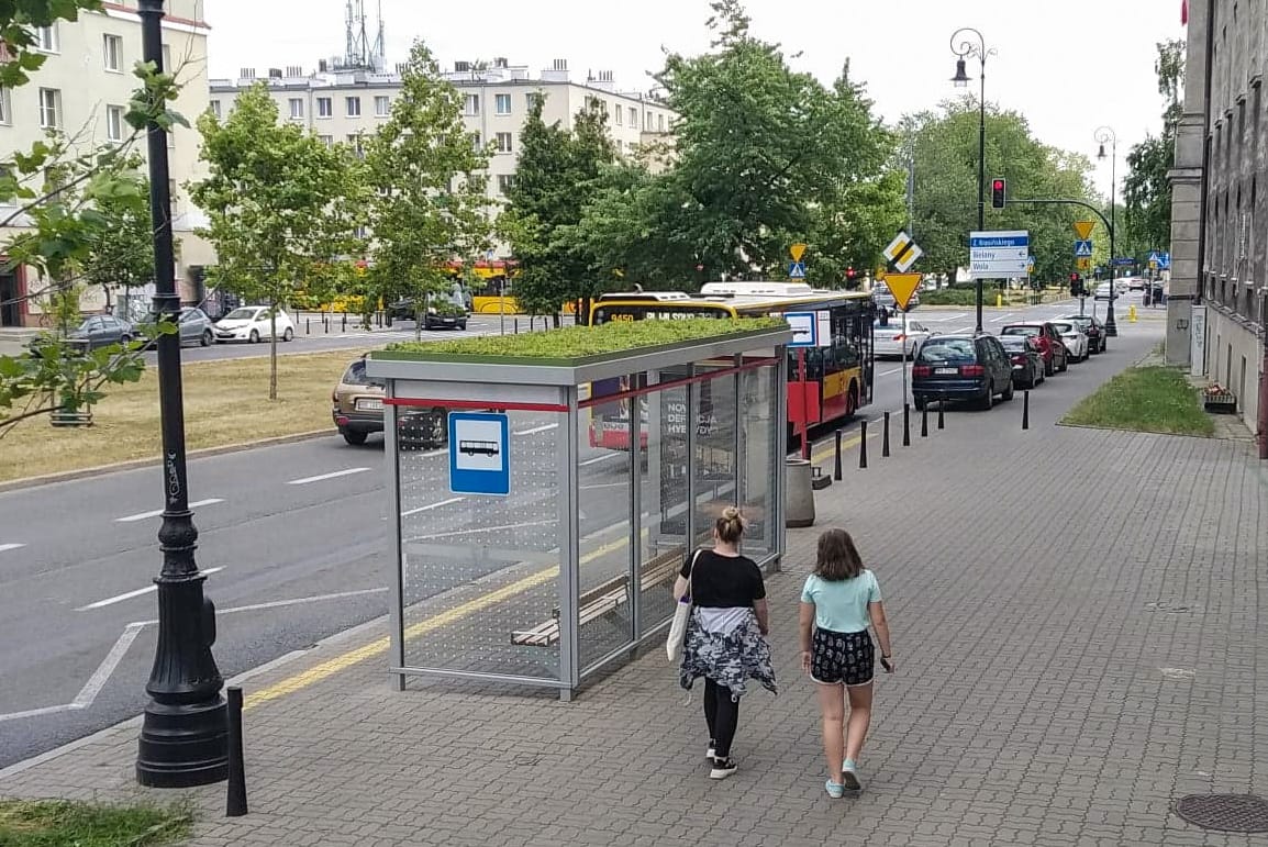Dwie dziewczynki przechodzą obok nowoczesnego przystanku autobusowego z zielonym dachem.
