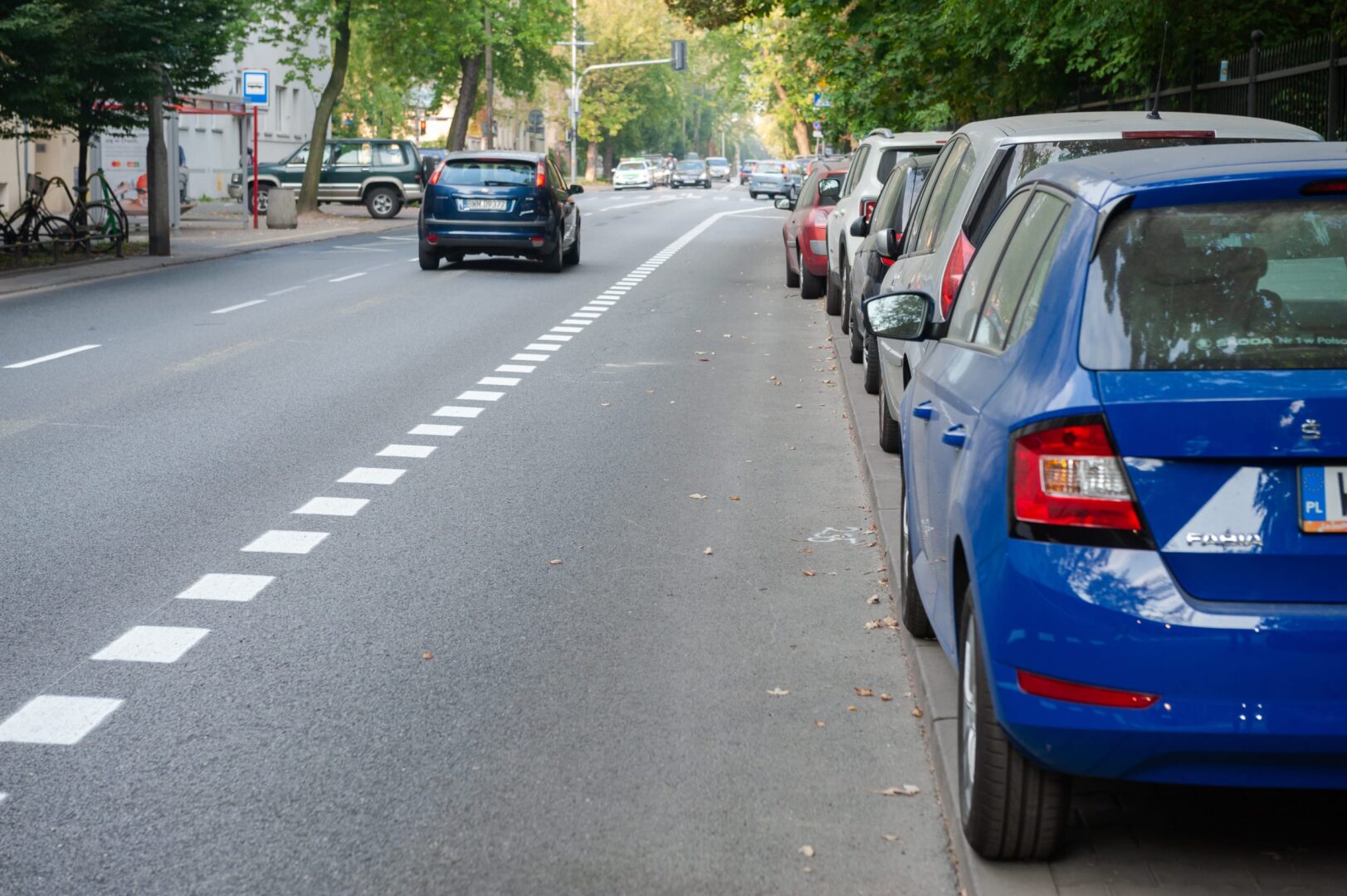 Samochody zaparkowane wzdłuż miejskiej ulicy, z pasem rowerowym oznaczonym białymi kwadratami.