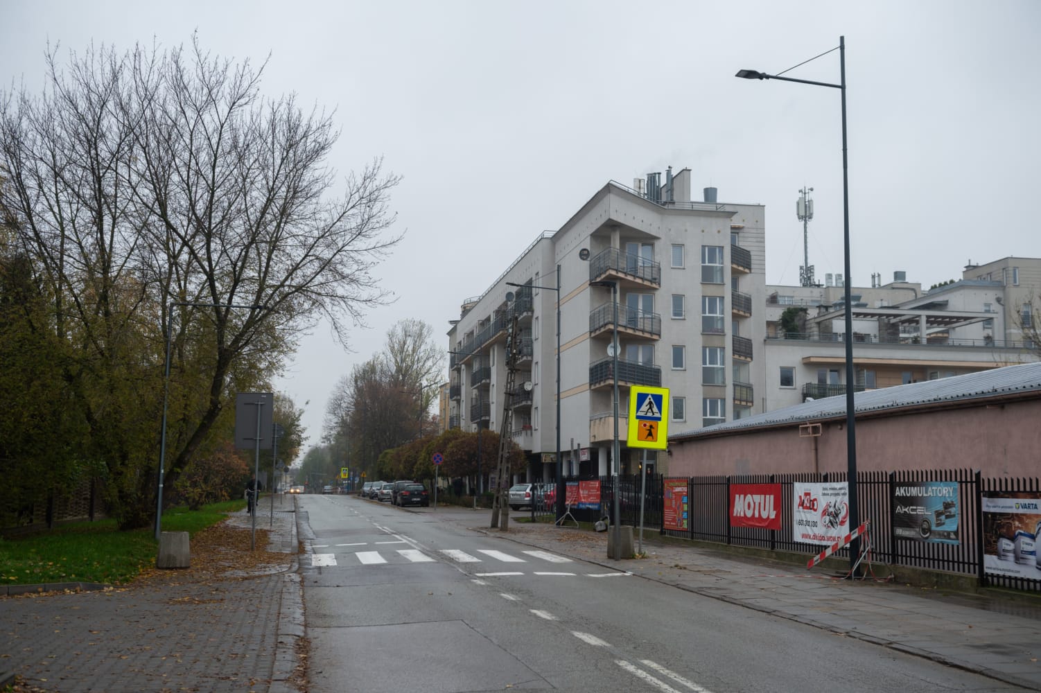 Scena ulicy mieszkalnej w Polsce z budynkami mieszkalnymi i przejściem dla pieszych. Widoczne reklamy Motul i Akcel.