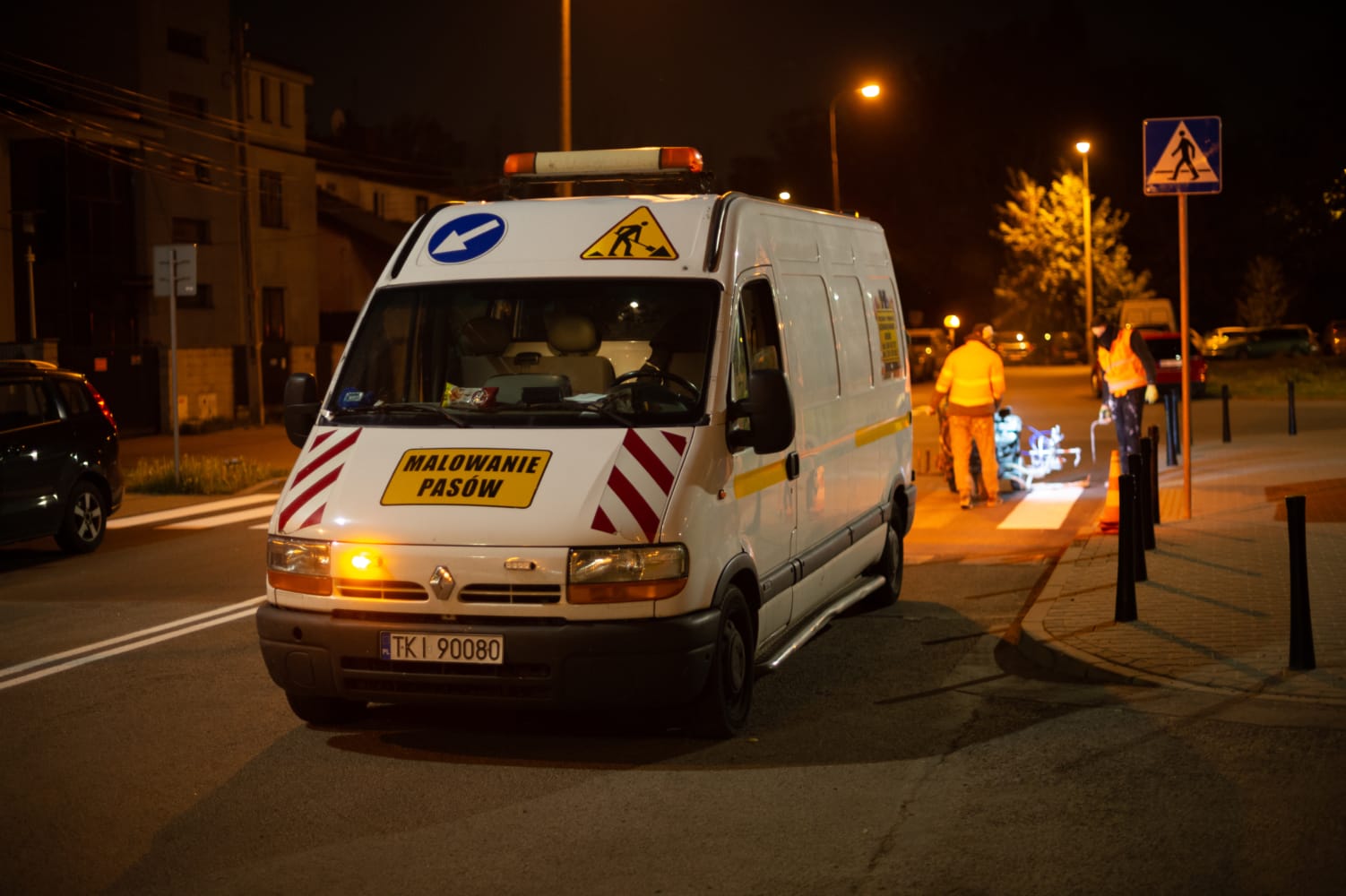 Oznakowanie dróg w nocy: Pracownicy malują linie przejść dla pieszych za pomocą maszyny do znakowania dróg. Na furgonetce widać „Malowanie Pasów”.