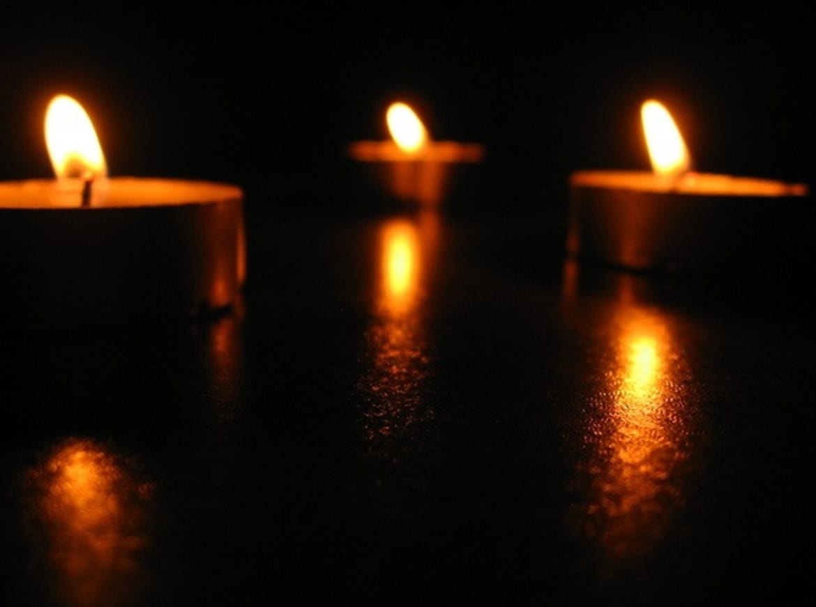 Trzy zapalone świeczki typu tealight świecą w ciemności, odbijając się od ciemnej powierzchni.