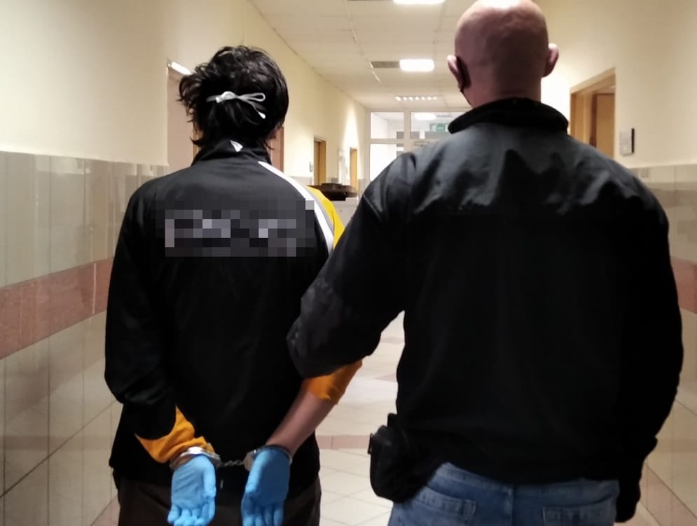 Policjant eskortujący podejrzanego w kajdankach korytarzem