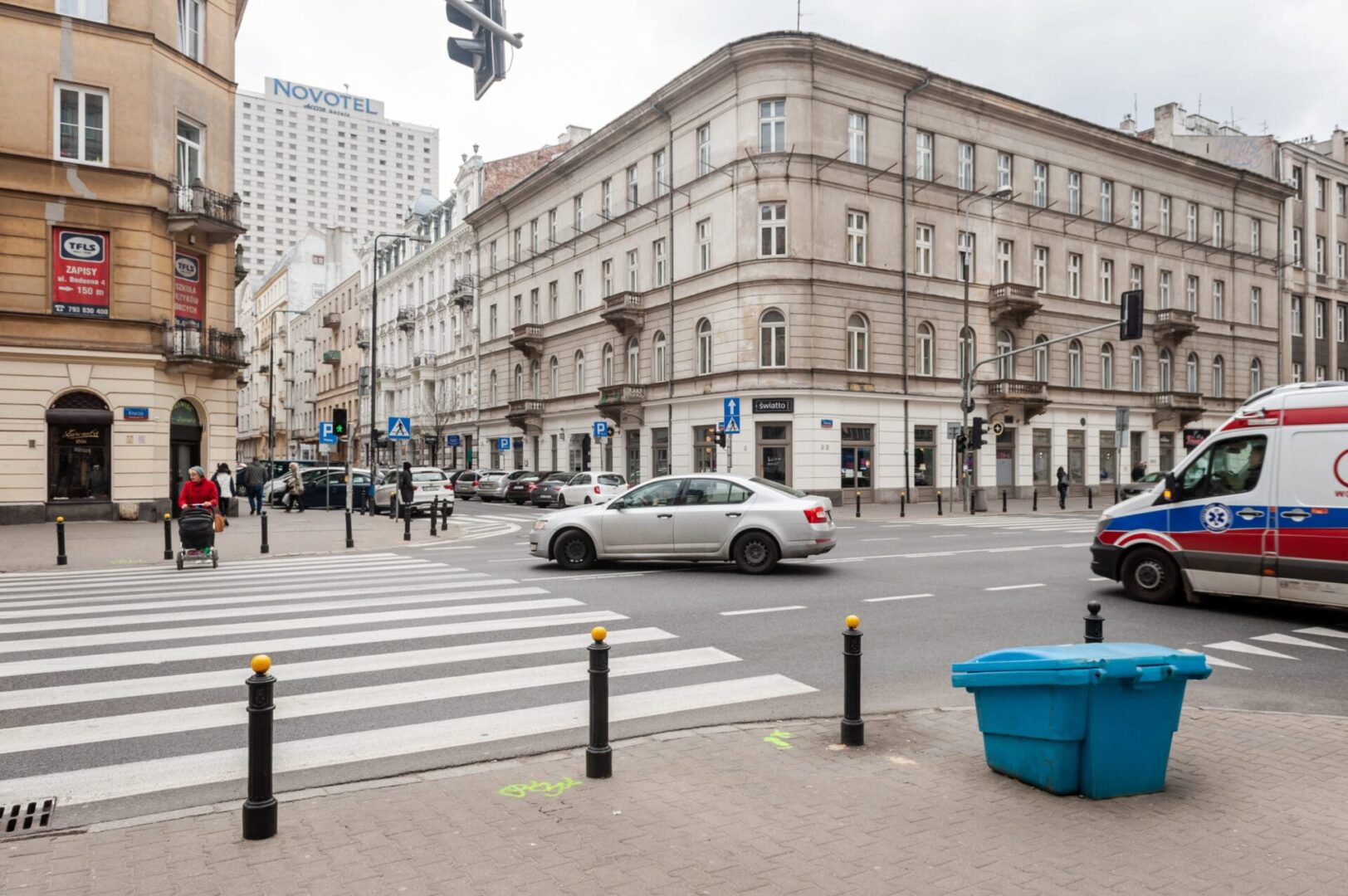 Scena uliczna w Warszawie z przejściem dla pieszych, samochodami i karetką pogotowia.