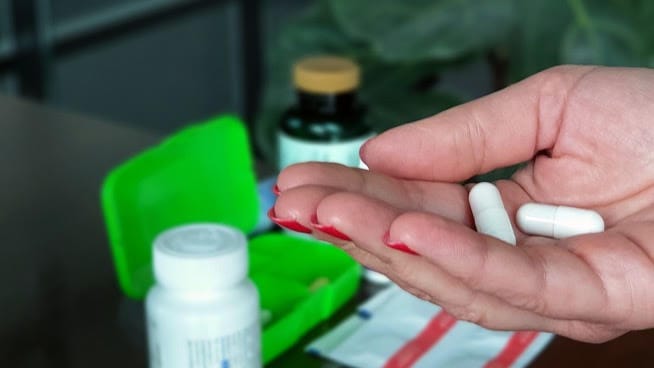 Ręka trzymająca dwie białe tabletki, w tle zielony organizer na tabletki i buteleczki z suplementami.