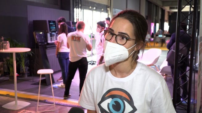 Kobieta w masce i okularach przemawia na konferencji technologicznej. Na jej koszulce jest logo z okiem.