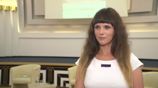 Młoda kobieta o długich brązowych włosach, ubrana w białą sukienkę, udziela wywiadu.