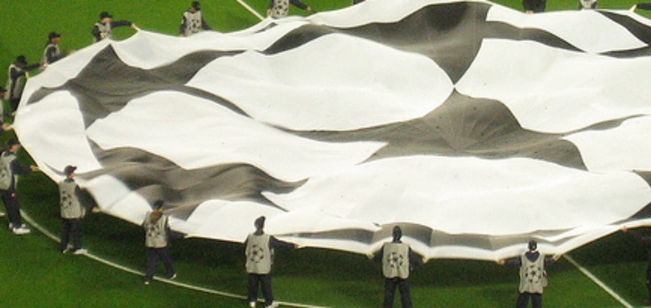 Duża flaga finału Ligi Mistrzów rozwijana przez liczne grono osób na boisku piłkarskim.