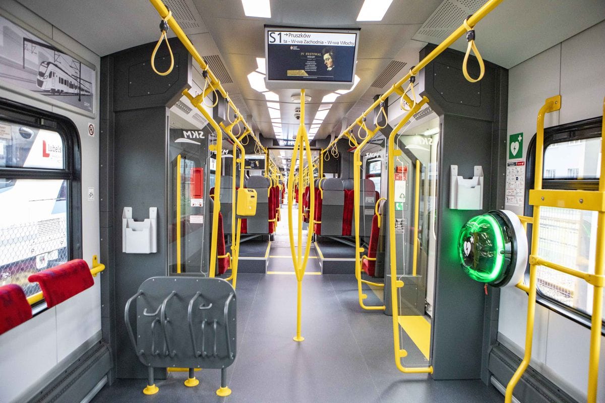 Wnętrze nowoczesnego pociągu podmiejskiego z żółtymi poręczami i czerwonymi siedzeniami. Znak docelowy pokazuje „Pruszków”.