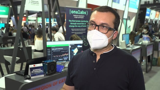 Mężczyzna w masce na twarzy prezentujący technologię monitorowania mleczanu na konferencji poświęconej technologiom medycznym. Widoczny baner „onalabs”.